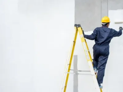 Ouvrier sur une échelle peignant un mur intérieur blanc.